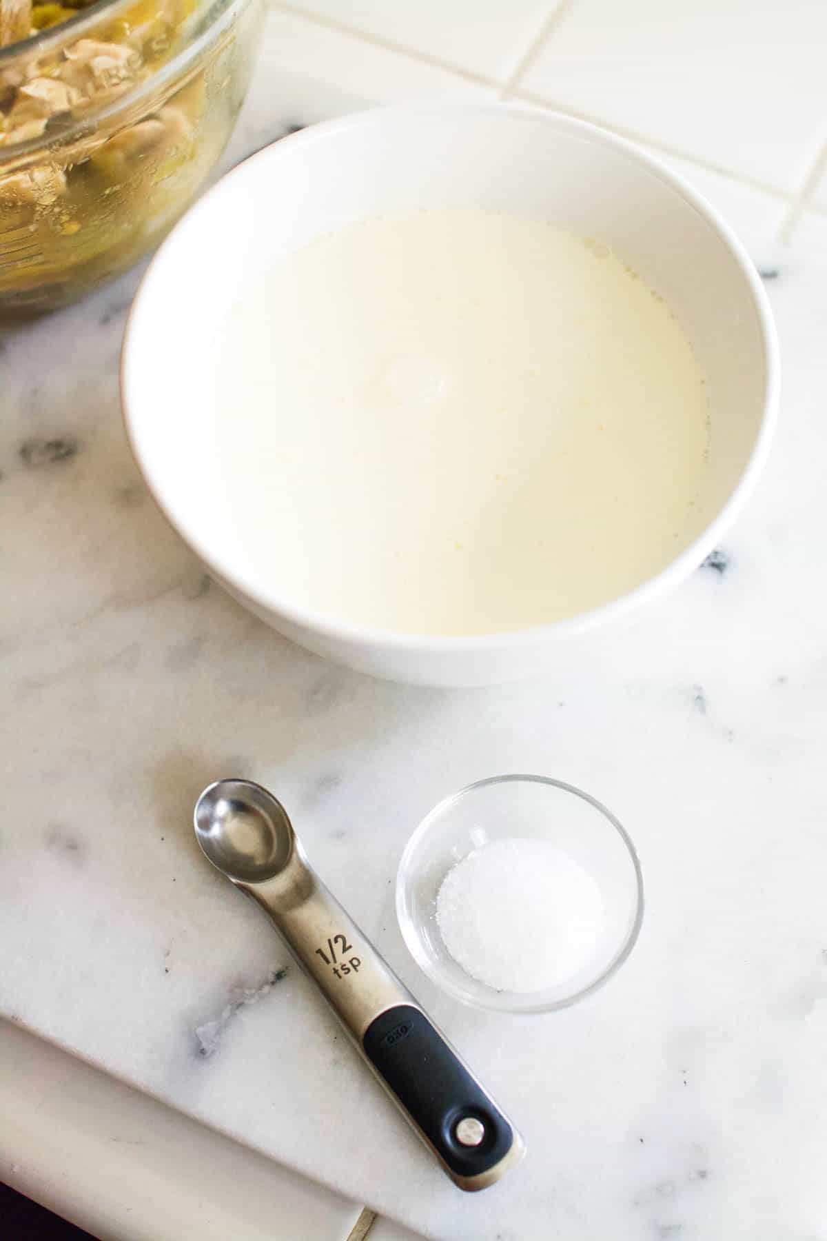 A bowl with whipping cream next to a measuring spoon and salt on a counter.