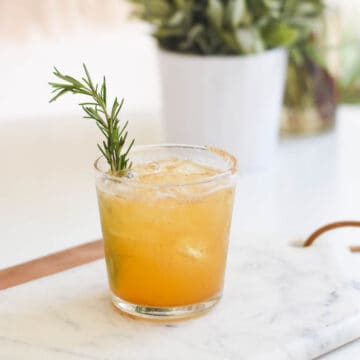 Cocktail in a glass on a table with a rosemary sprig.