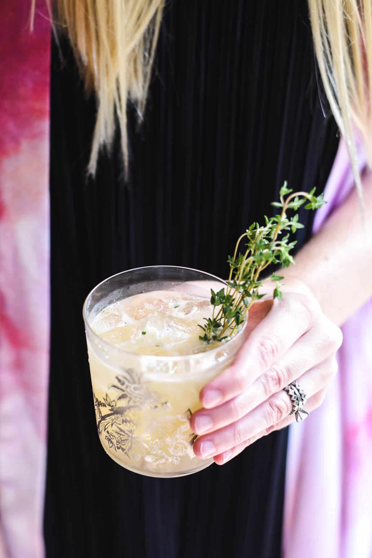 Woman holding a cocktail with a sprig of fresh thyme.