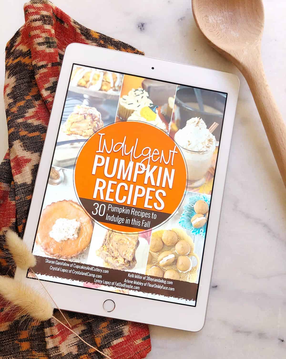 Ipad on a table with a digital pumpkin cookbook on it lying on a cutting board next to a wooden spoon.