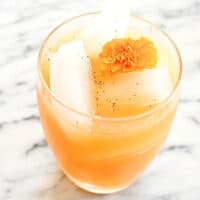 A pear cocktail in a glass on a marble table with an edible marigold on top.