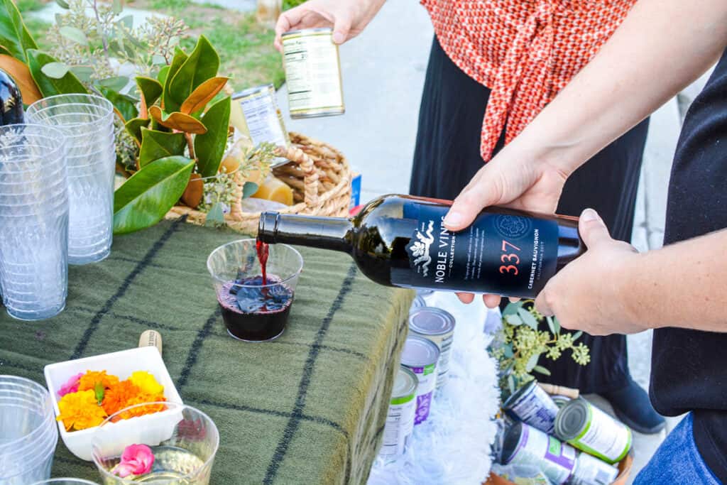 Woman pouring a glass of red wine with a lady putting canned food in a basket.