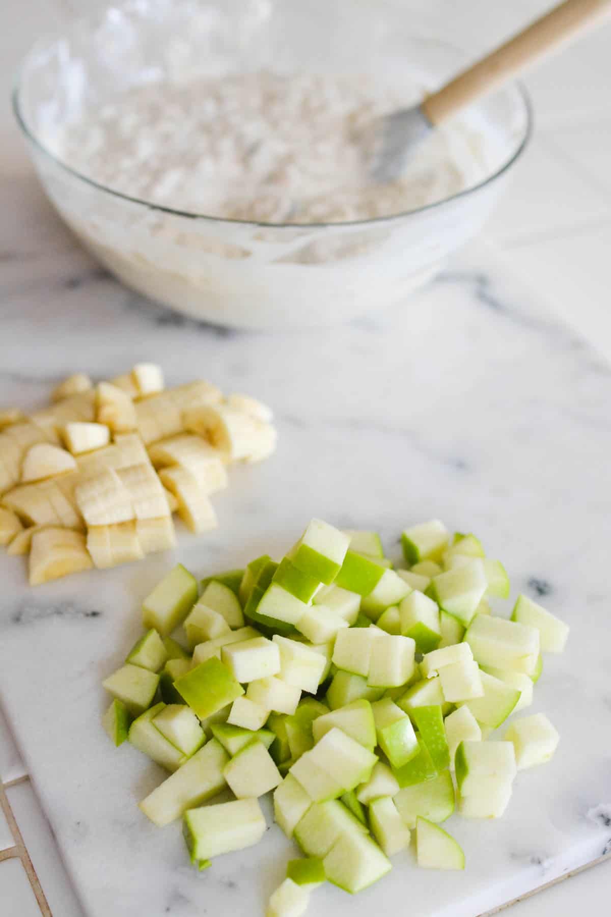 Bowl of yogurt and oats in the background on a cutting board with chopped green apple and bananas in front of it.