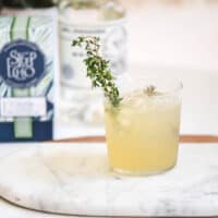 Close up of cocktail cup with fresh thyme garnish in it on a marble cutting board.