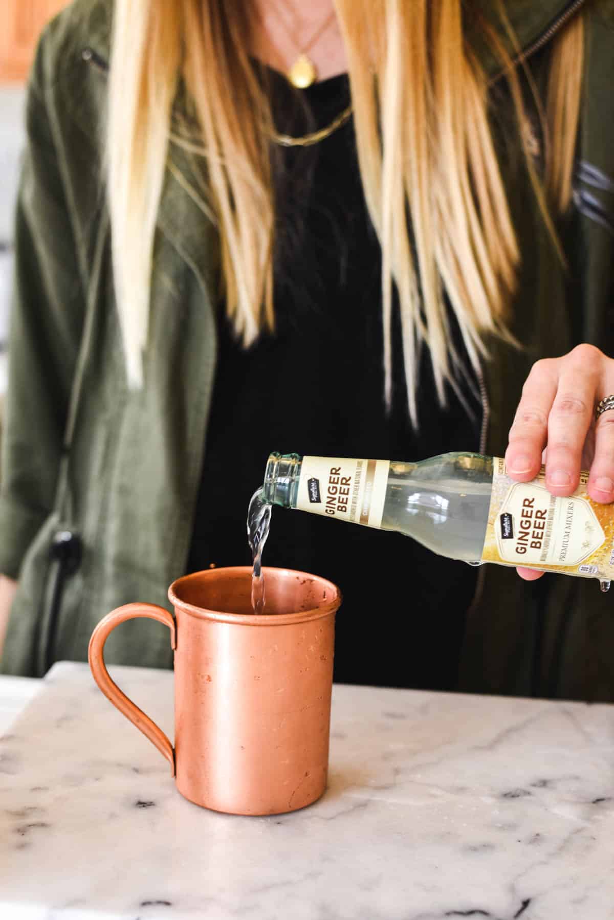Woman adding ginger beer to a copper Moscow Mule mug.