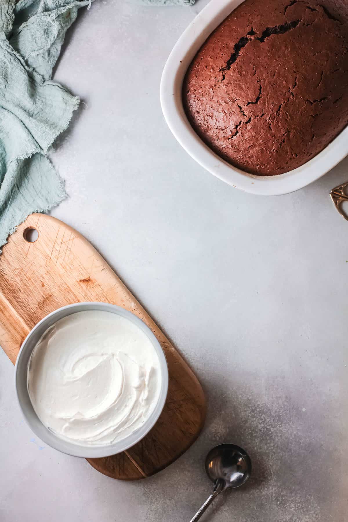 Frosting in a bowl on a table next to a chocolate cake.