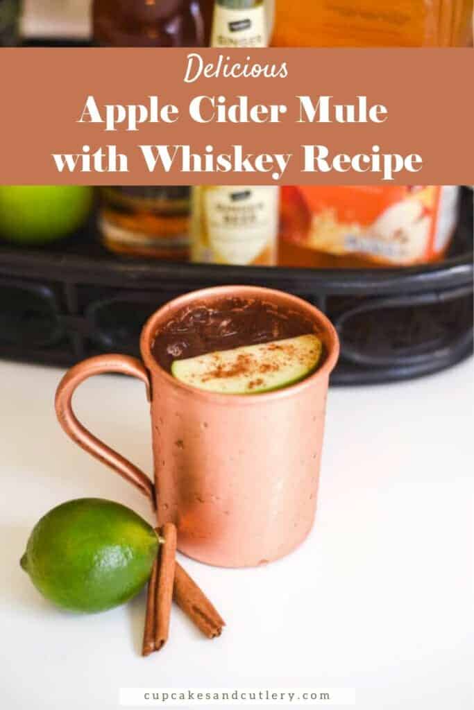 Text: Apple Cider Mule with Whiskey Recipe with a copper mug on a table topped with a slice of green apple dusted with pie spice and lime and cinnamon sticks on the table next to it.