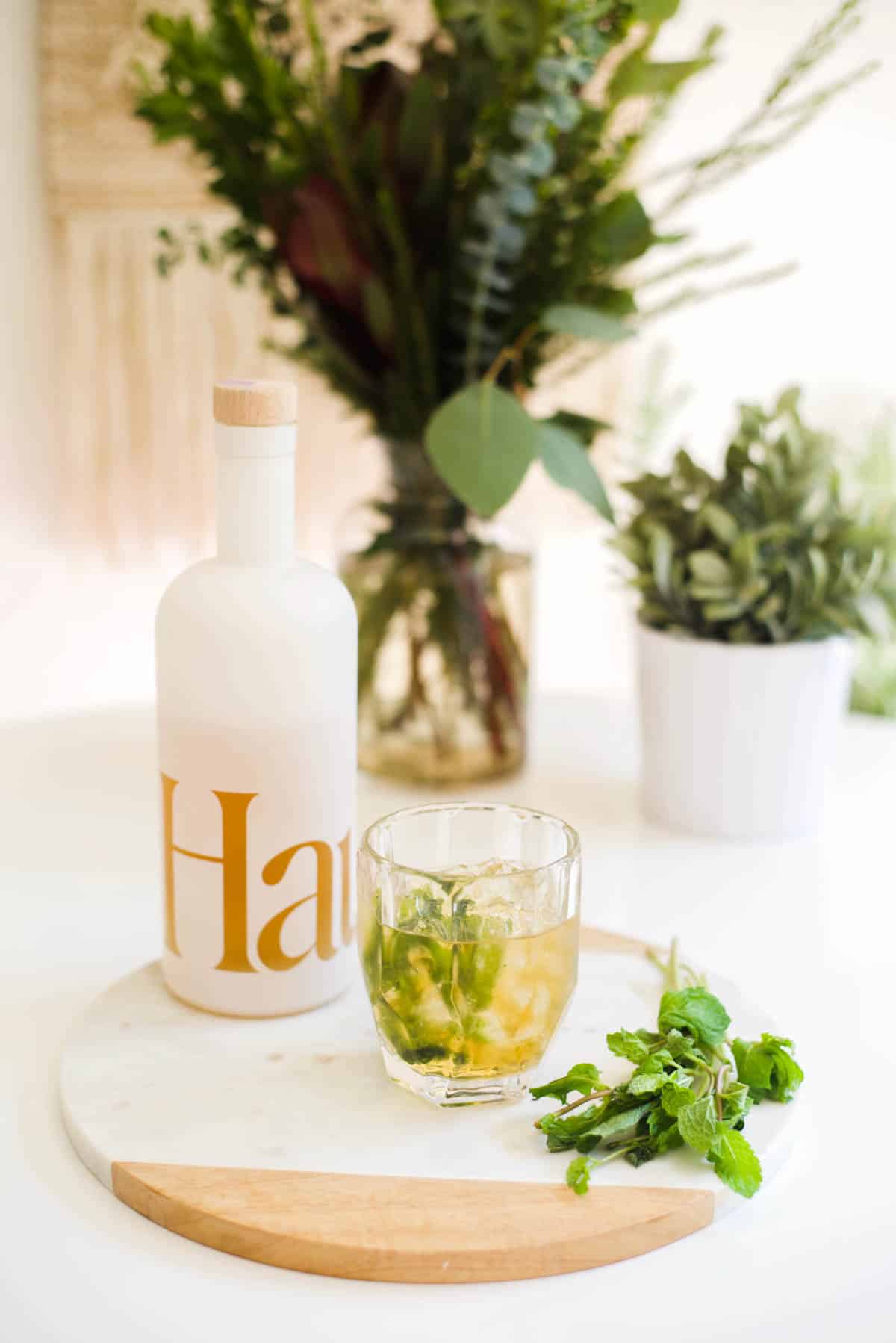 A cocktail on a small try next to a bunch of fresh mint next to a bottle of Haus wine.