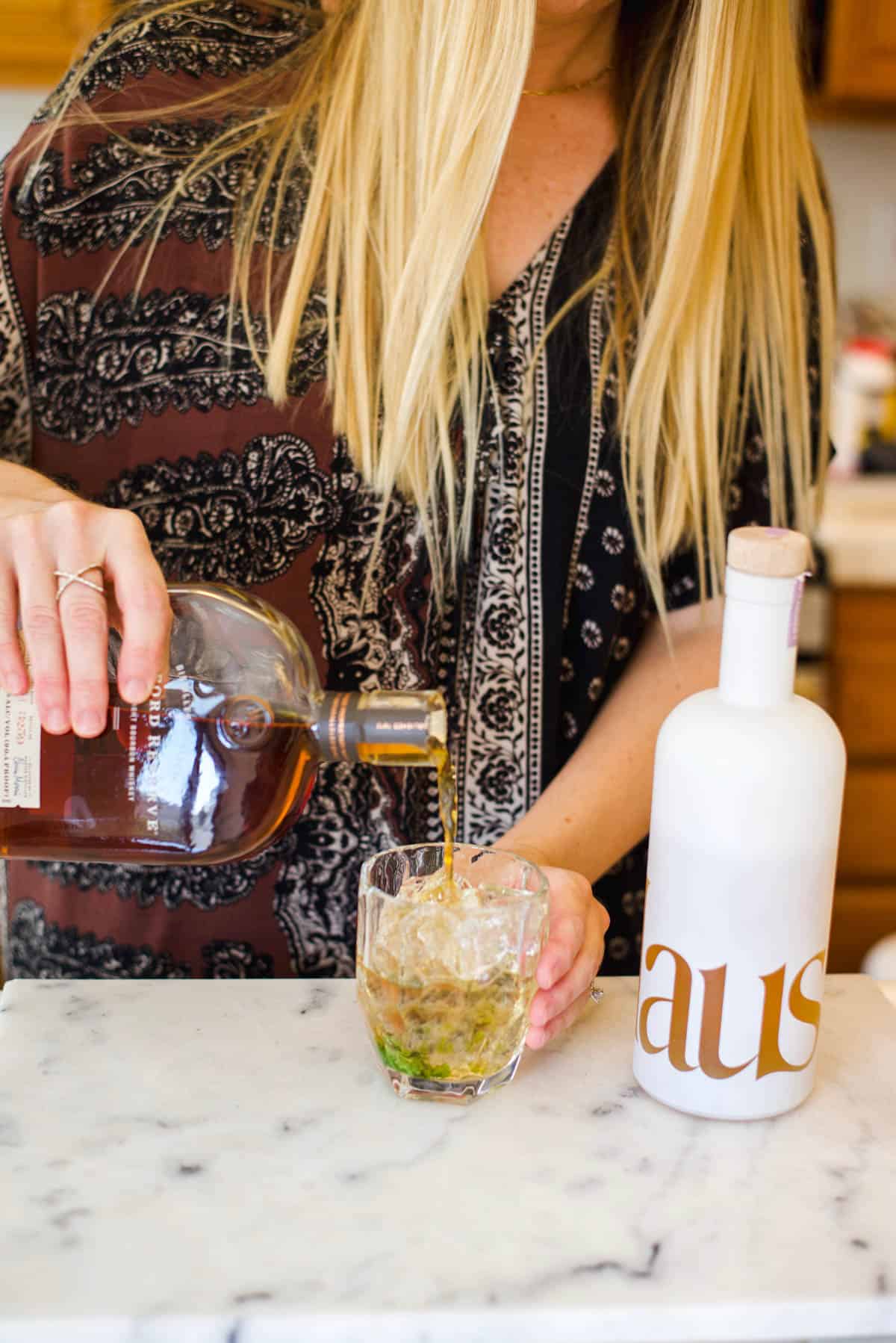 Woman pouring bourbon into a cocktail glass on a counter.
