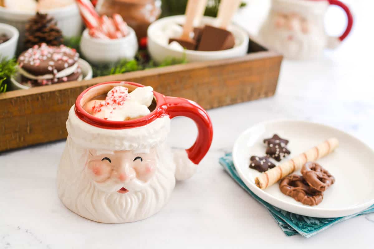 Close up of a Santa coffee mug on a table next to a small plate of cookies.