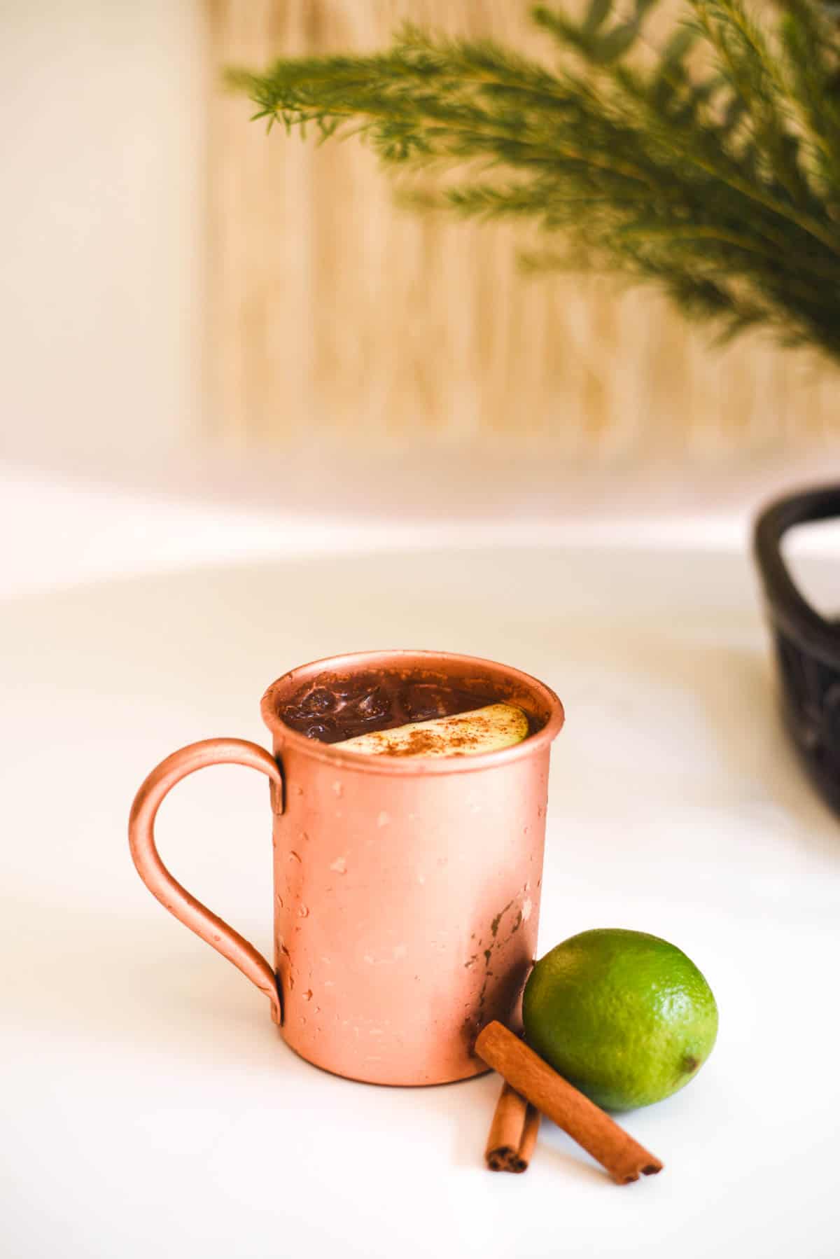 A copper mug on a table with a lime and cinnamon sticks sitting next to it.