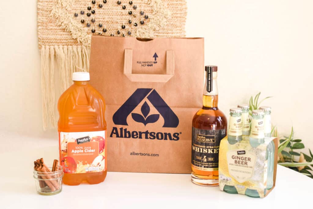 An Albertsons bag on a table next to a bottle of apple cider, a bottle of whiskey and a pack of ginger beer.