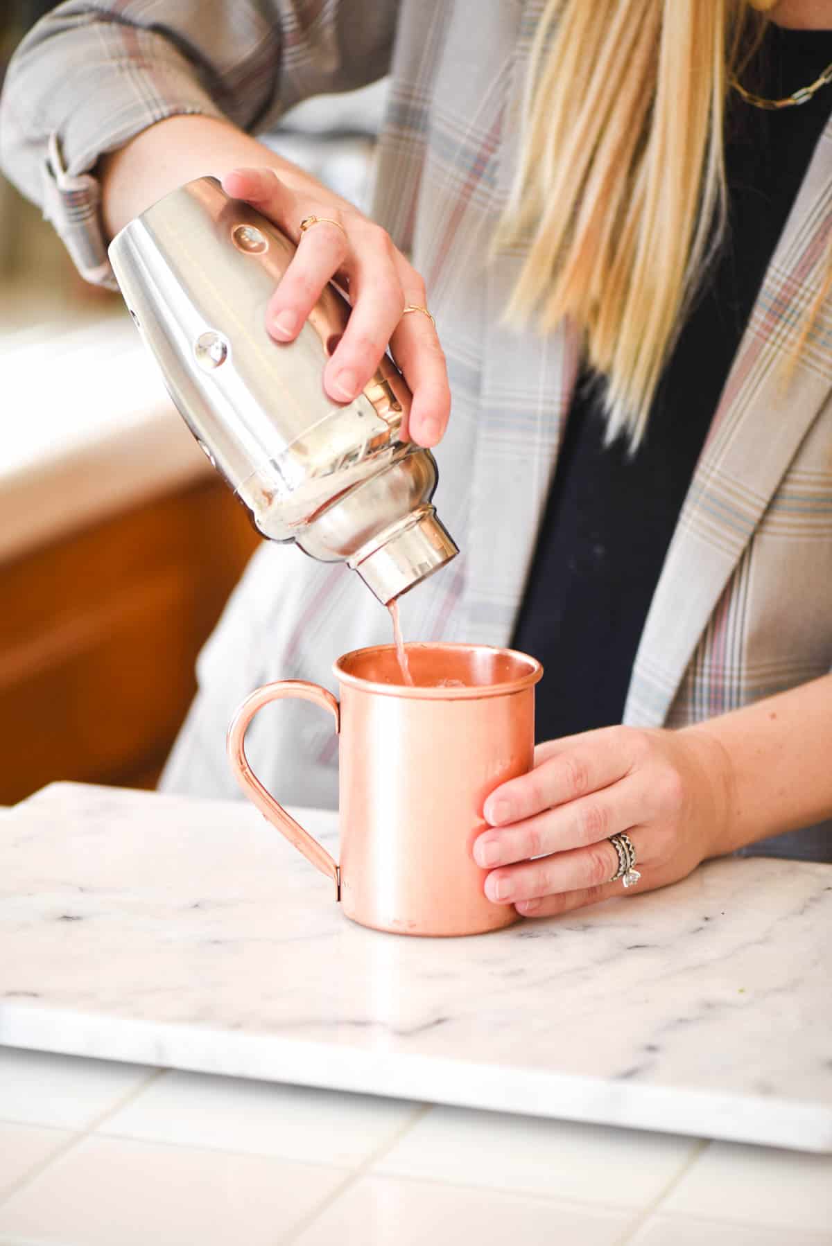 Woman pouring a cocktail from a cocktail shaker into a copper mug.