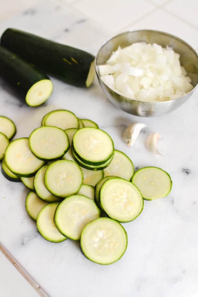 Sliced zucchini on a cutting board next to a bowl of chopped onions and a few garlic cloves.