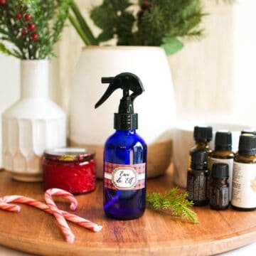Square image of a wooden platter with a glass bottle filled with holiday room spray, essential oils, greenery, and candy canes.