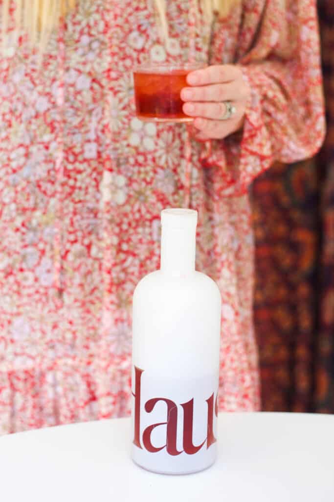 Woman holding a glass of rose with a bottle on the table in front of her.