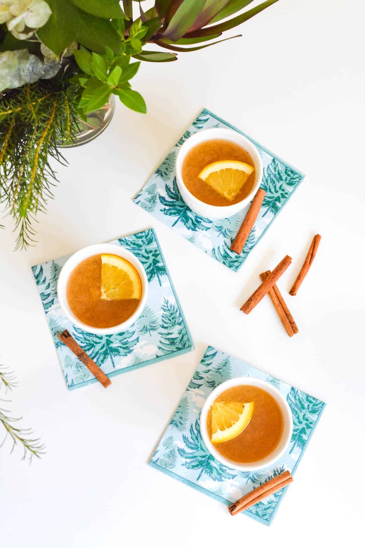 Overhead shot of small white mugs holding hot apple cider on a table with napkins and cinnamon sticks.