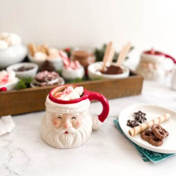 A mug of hot cocoa in a Santa face mug next to a tray full of toppings for a drink station.