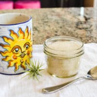 A small jar of lavender sugar on a counter next to a coffee mug with a sun on it.