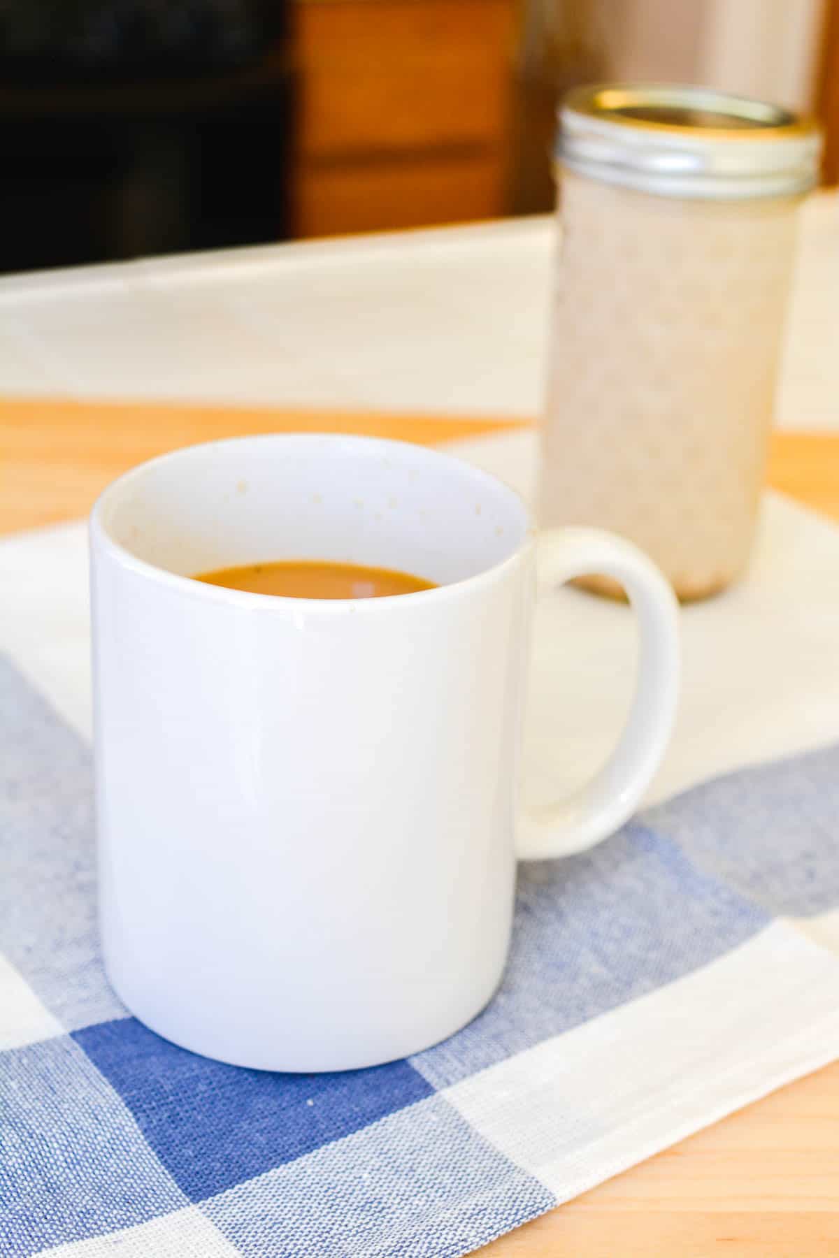 A coffee cup on a counter with a jar behind it holding cinnamon homemade coffee creamer.