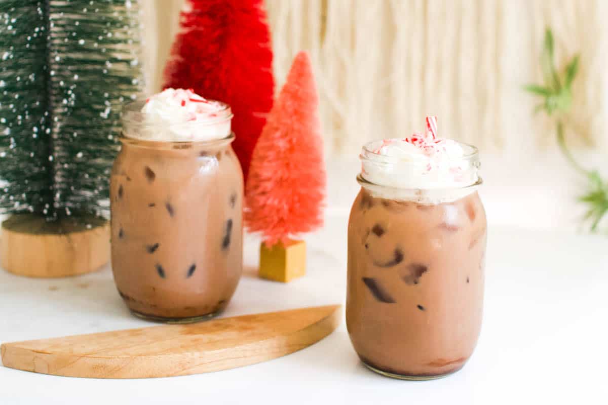 2 Old fashioned chocolate egg cream sodas in jars on a table topped with whipped cream and crushed candy canes with colorful Christmas tree decorations in the background. 