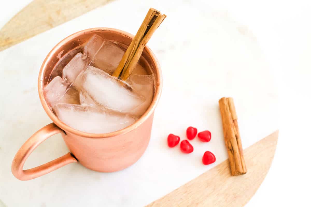 Overhead view of a copper cup holding a cocktail with cinnamon candies and a cinnamon stick on a table next to it.