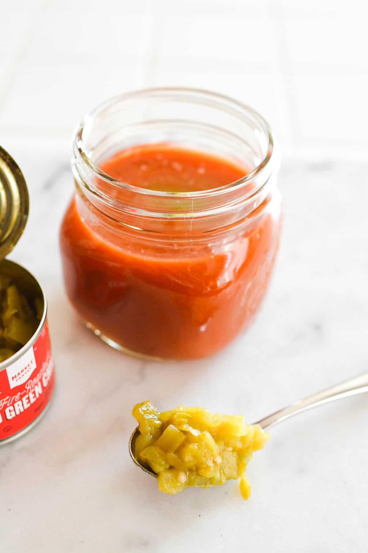 A jar full of tomato juice with a spoon holding green chiles.