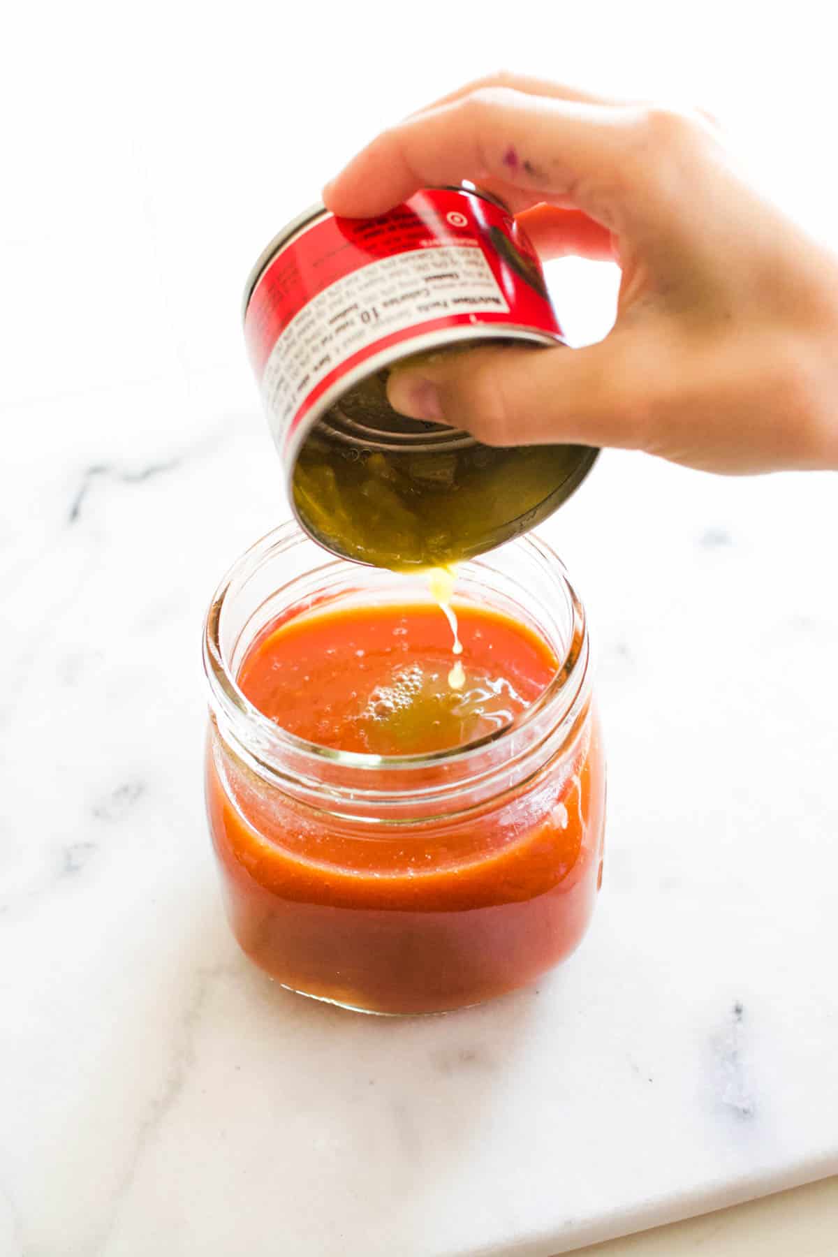 A jar of tomato juice with someone squeezing the juice from a can of green chiles into the juice.