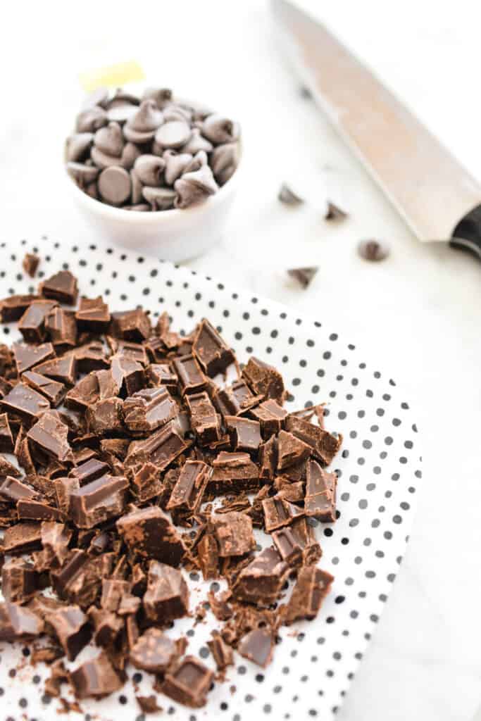Close up of a plate with chopped chocolate and a measuring cup with chocolate chips in the background. 