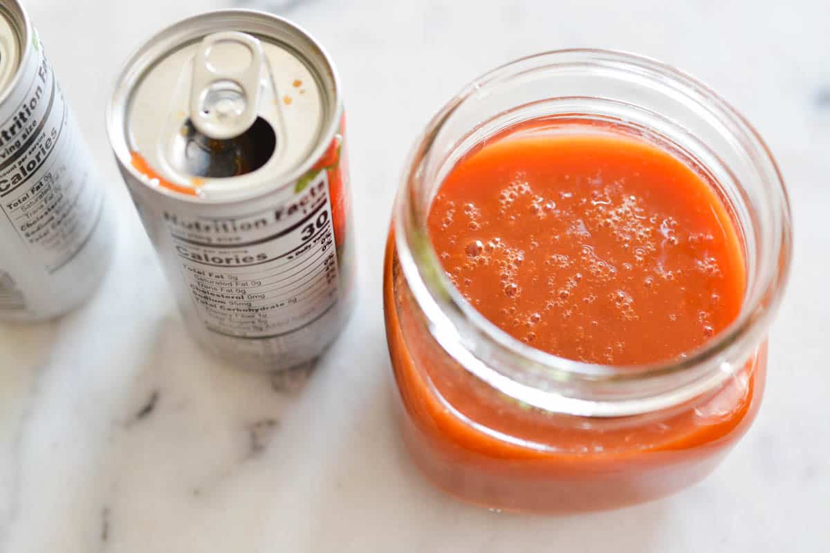Overhead view of a jar holding tomato juice next to an open can of V8.