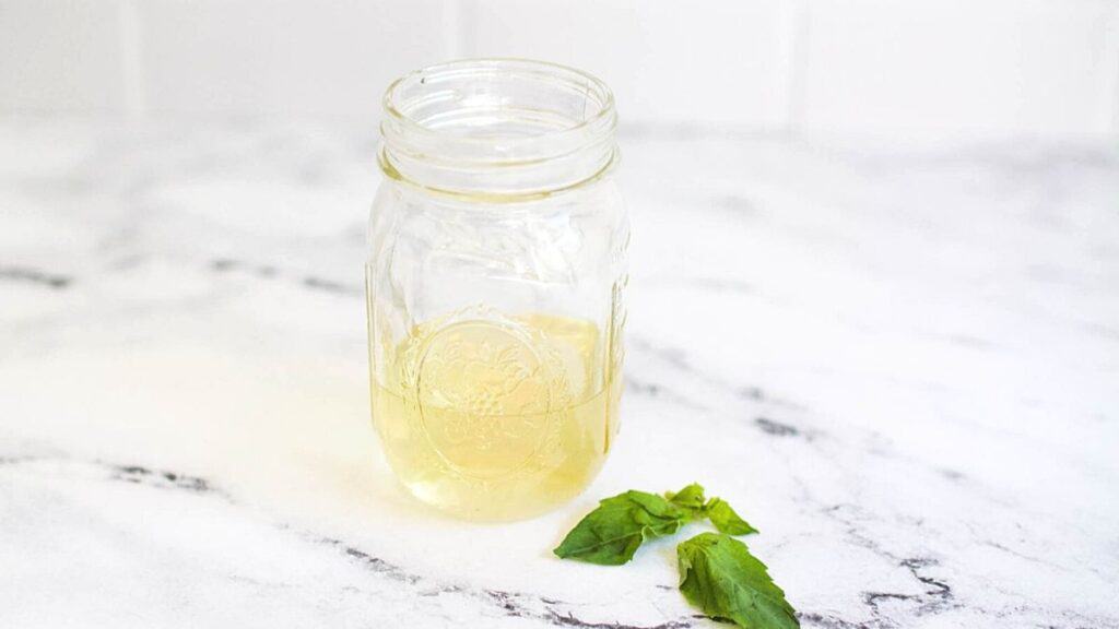 Close up of a jar with simple syrup on a counter with basil leaves to the right of it.
