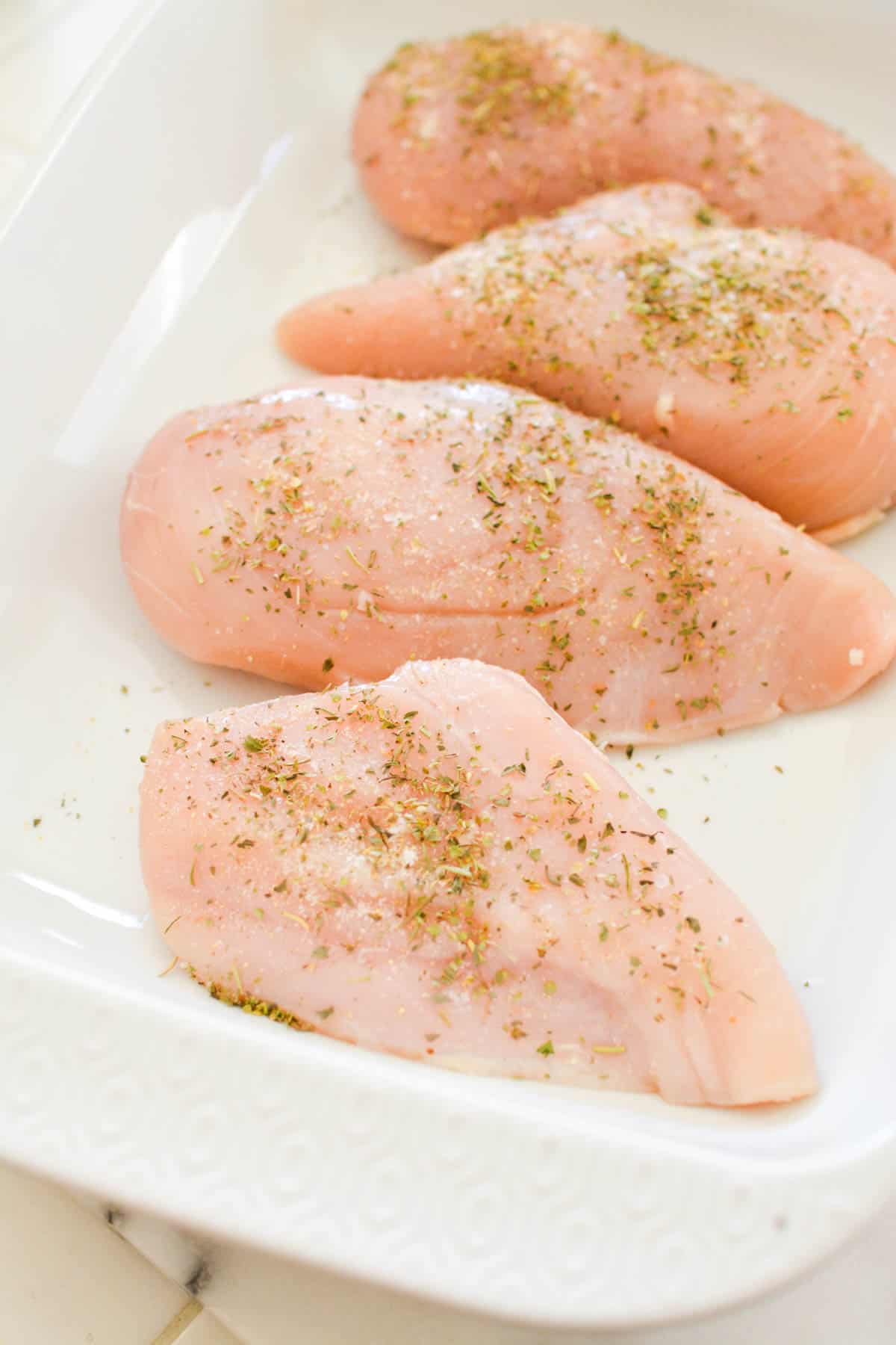 A baking dish with raw chicken breasts topped with Italian seasoning.