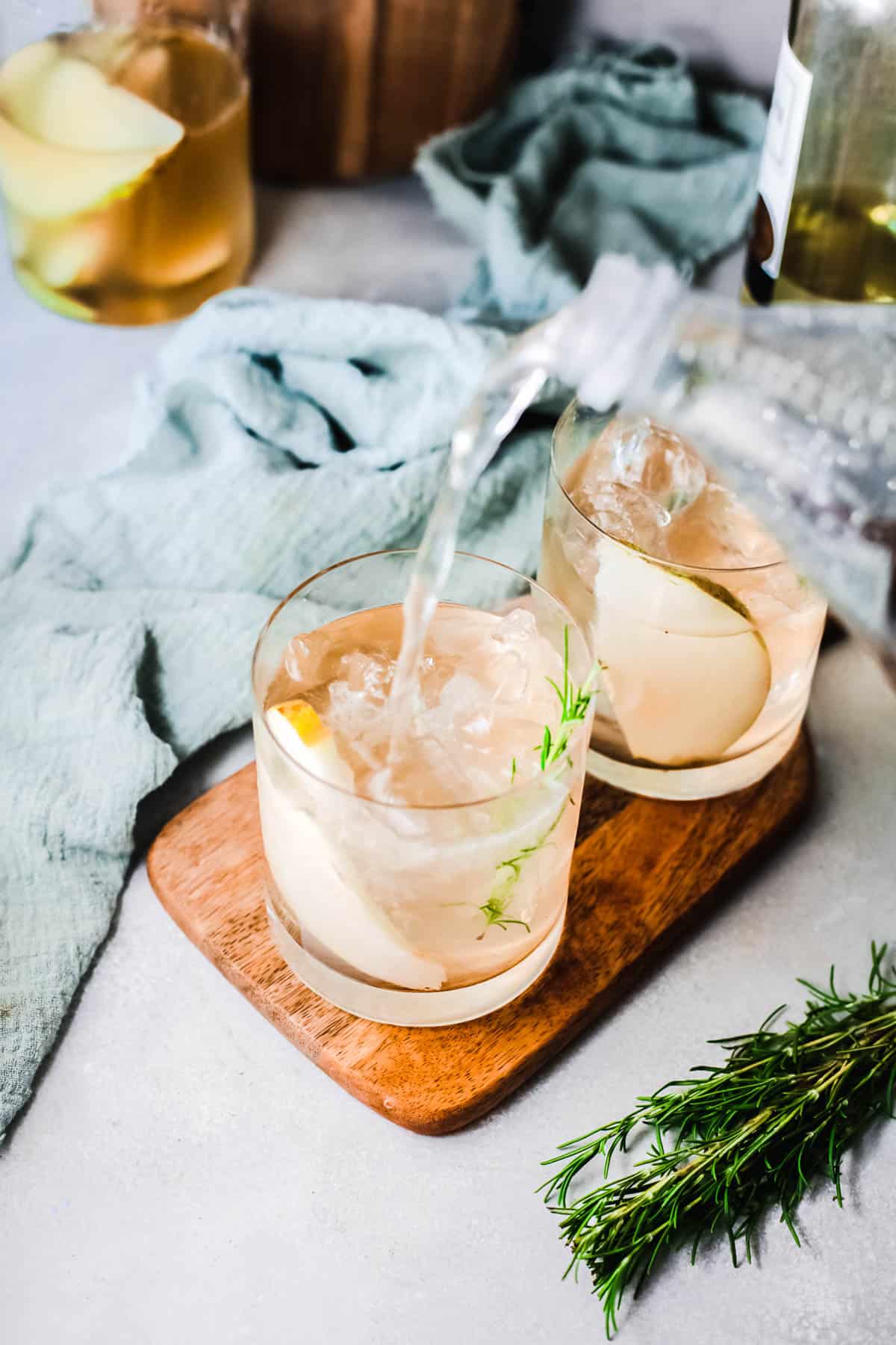2 cocktail glasses each with a slice of pear and rosemary sprig on a wood tray with club soda bing poured into one glass.