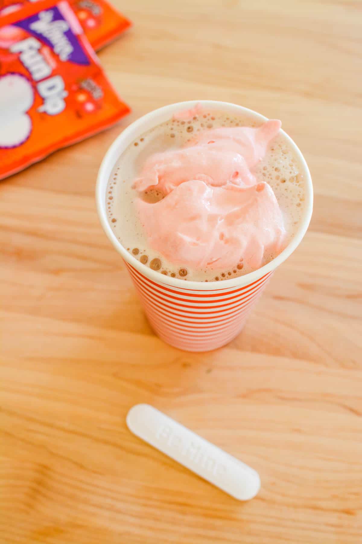 Cup of hot chocolate topped with pink whipped cream on a counter next to Fun Dip Candy.
