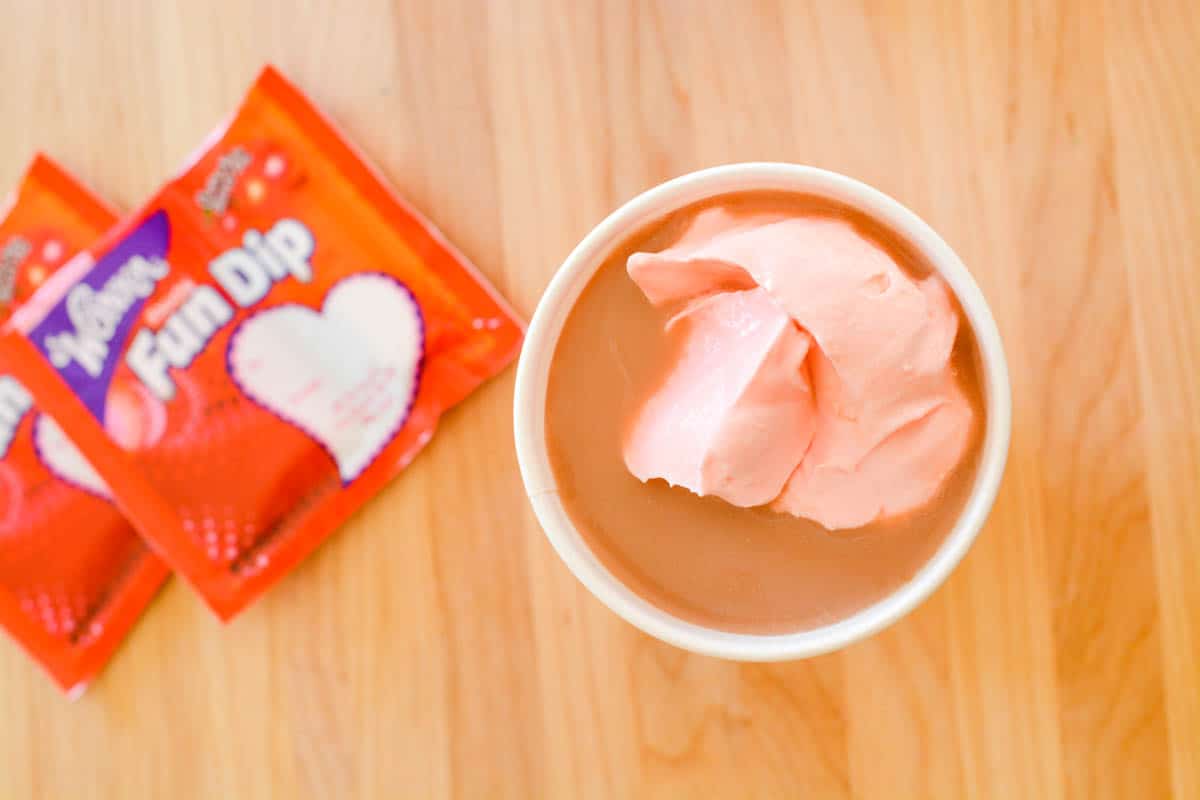 Overhead view of a cup with hot chocolate topped with pink whipped cream next to Fun Dip packets.