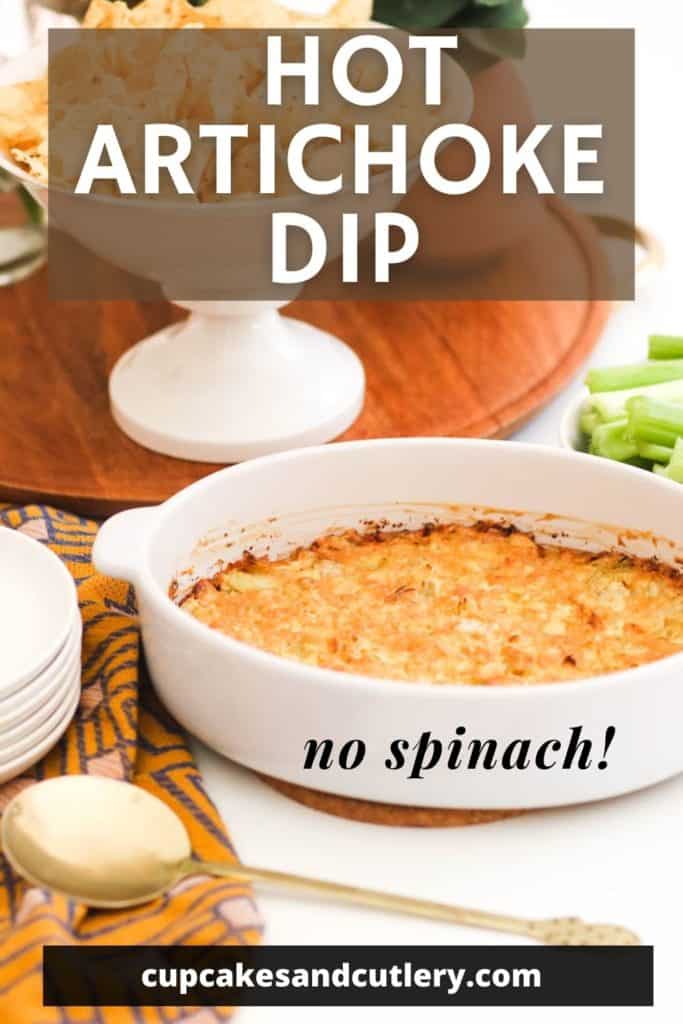 Close up of a white dish of baked arthcike dip on a table with serving plates and a white bowl fo chips in the background.
