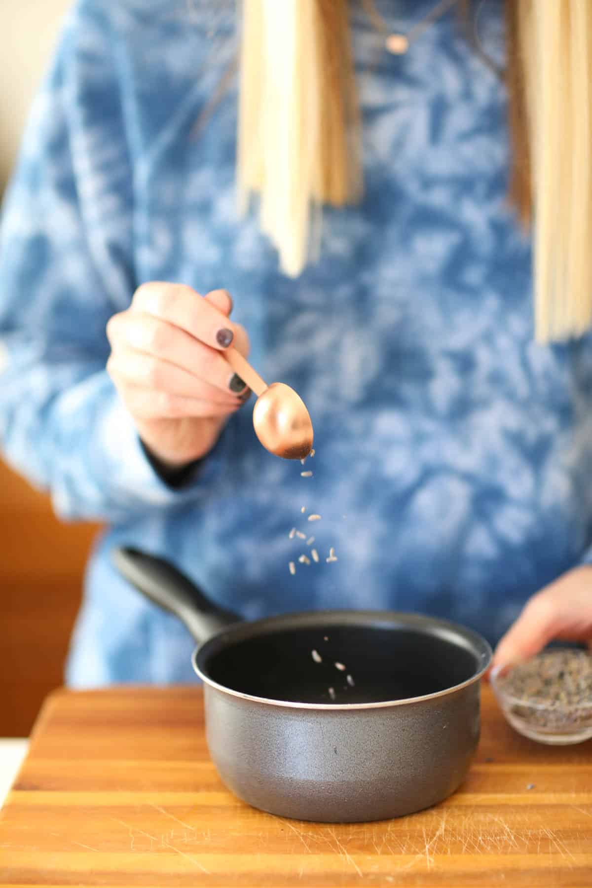 Woman adding lavender buds from a measuring spoon into a saucepan. 