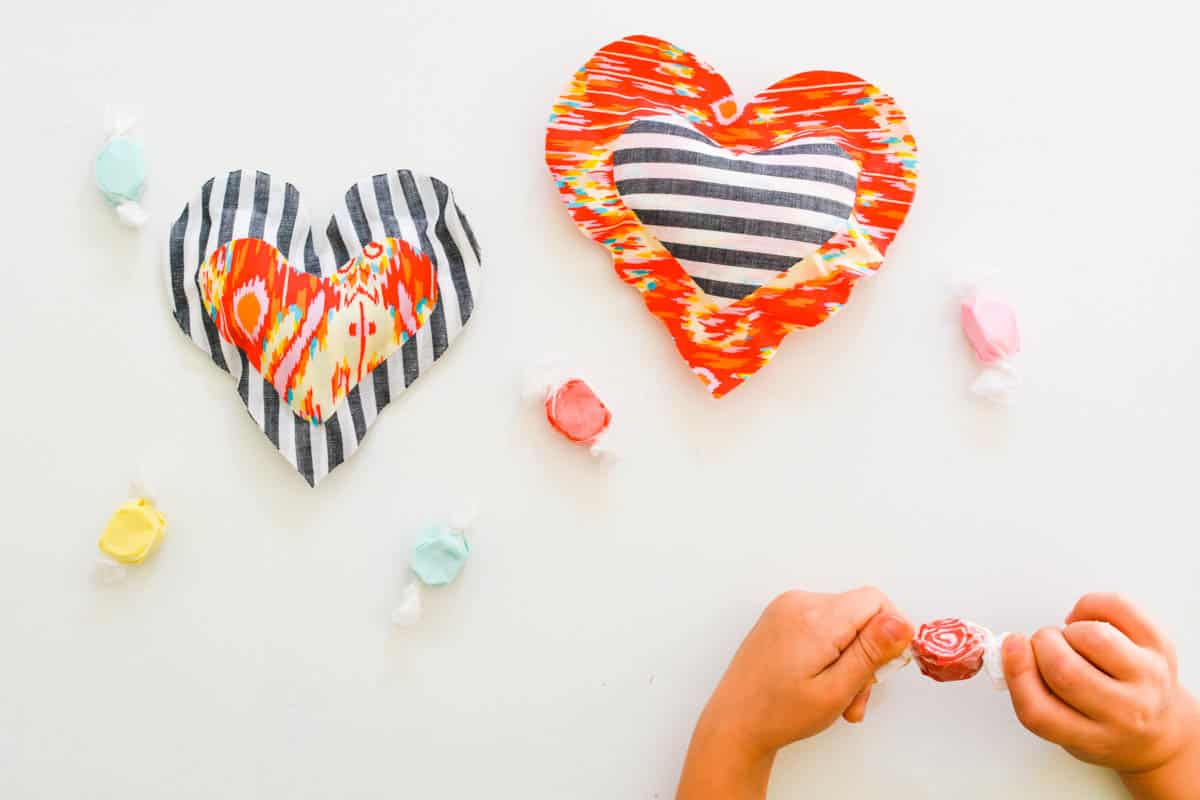 Small homemade heart shaped pillows on a table next to candy and a kid's hands unwrapping a piece of candy.