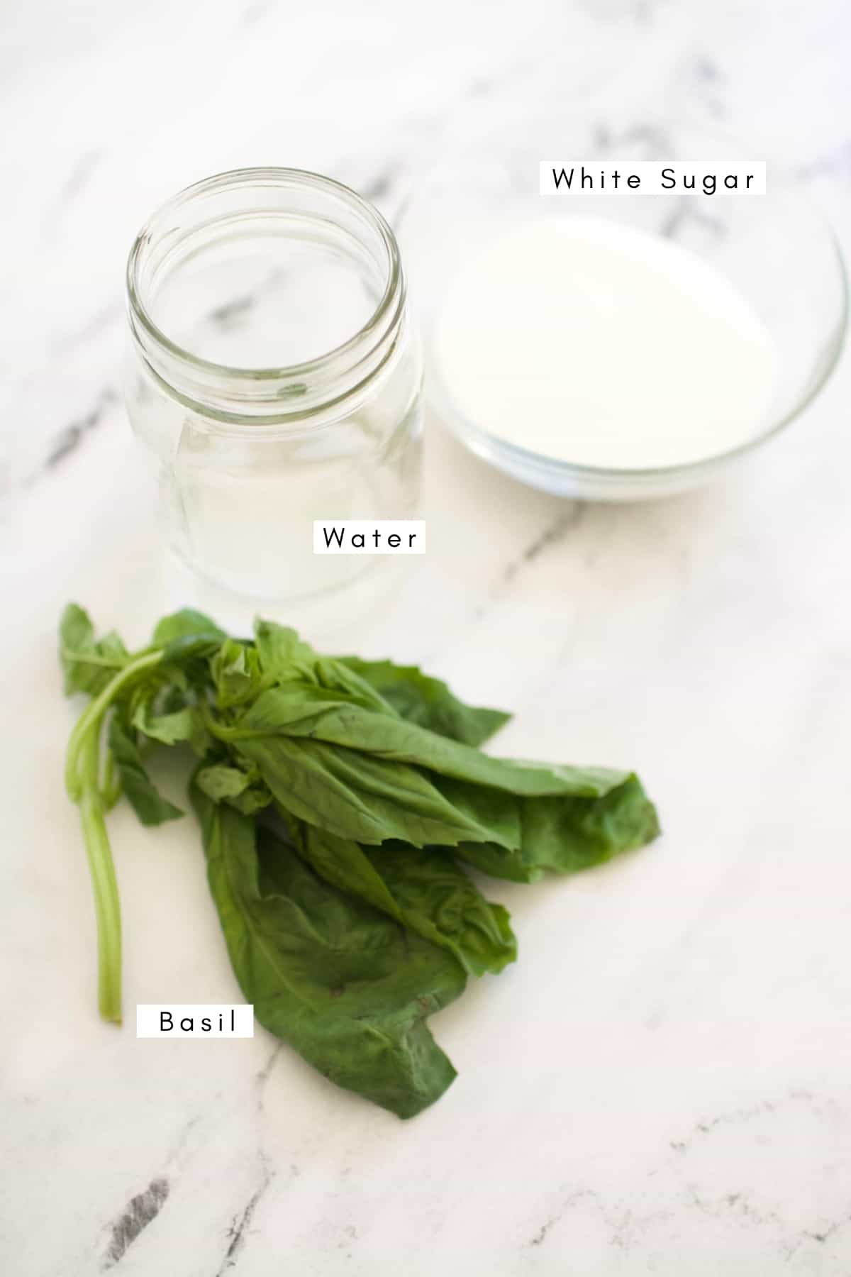 Fresh basil laying on a counter next to a jar of water and a small bowl with white sugar.