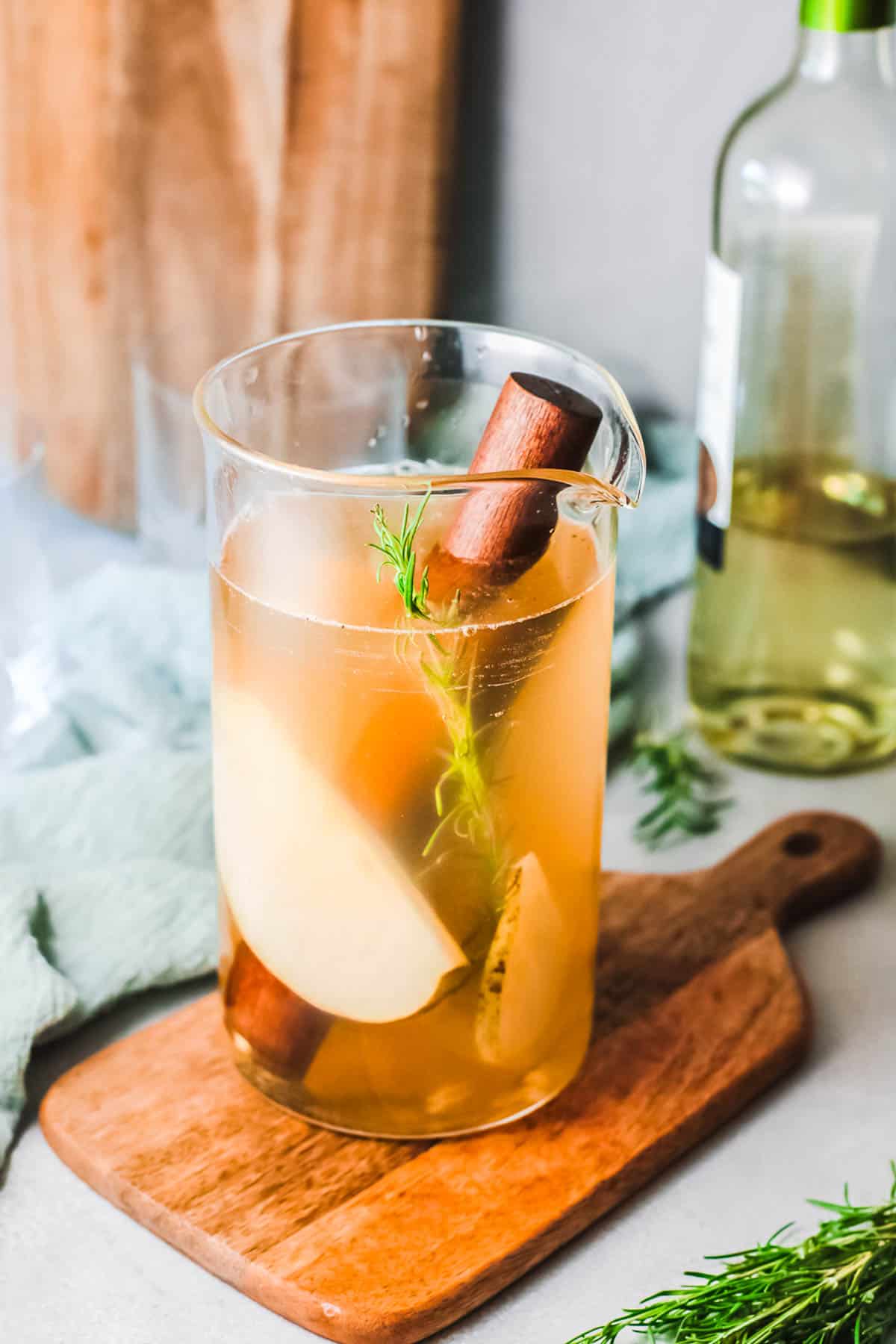 A glass pitcher with juices, wine, rosemary and sliced pears with a wooden muddler in it sitting on top of a wooden cutting board.