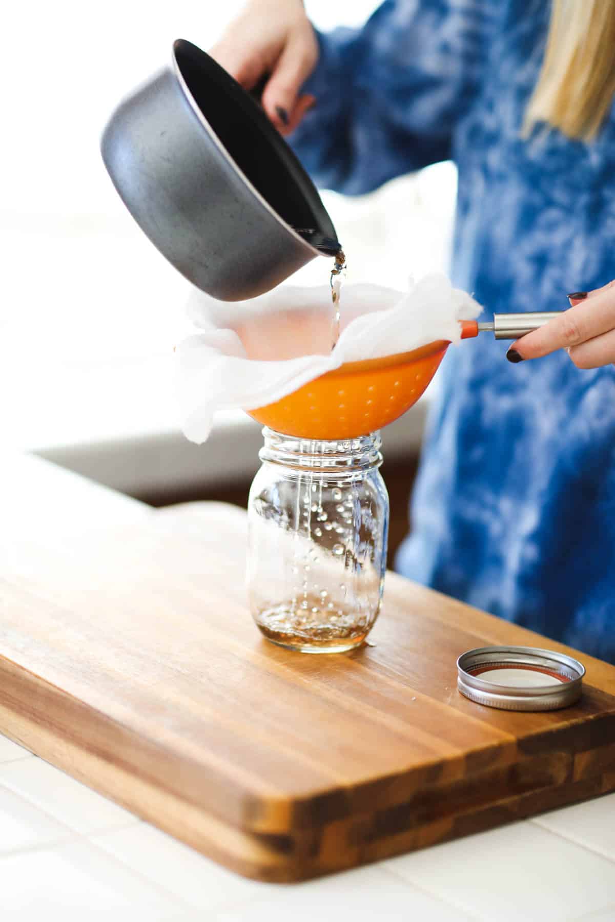 A woman pours tea from a saucepan through a cheesecloth lined strainer into a glass jar.