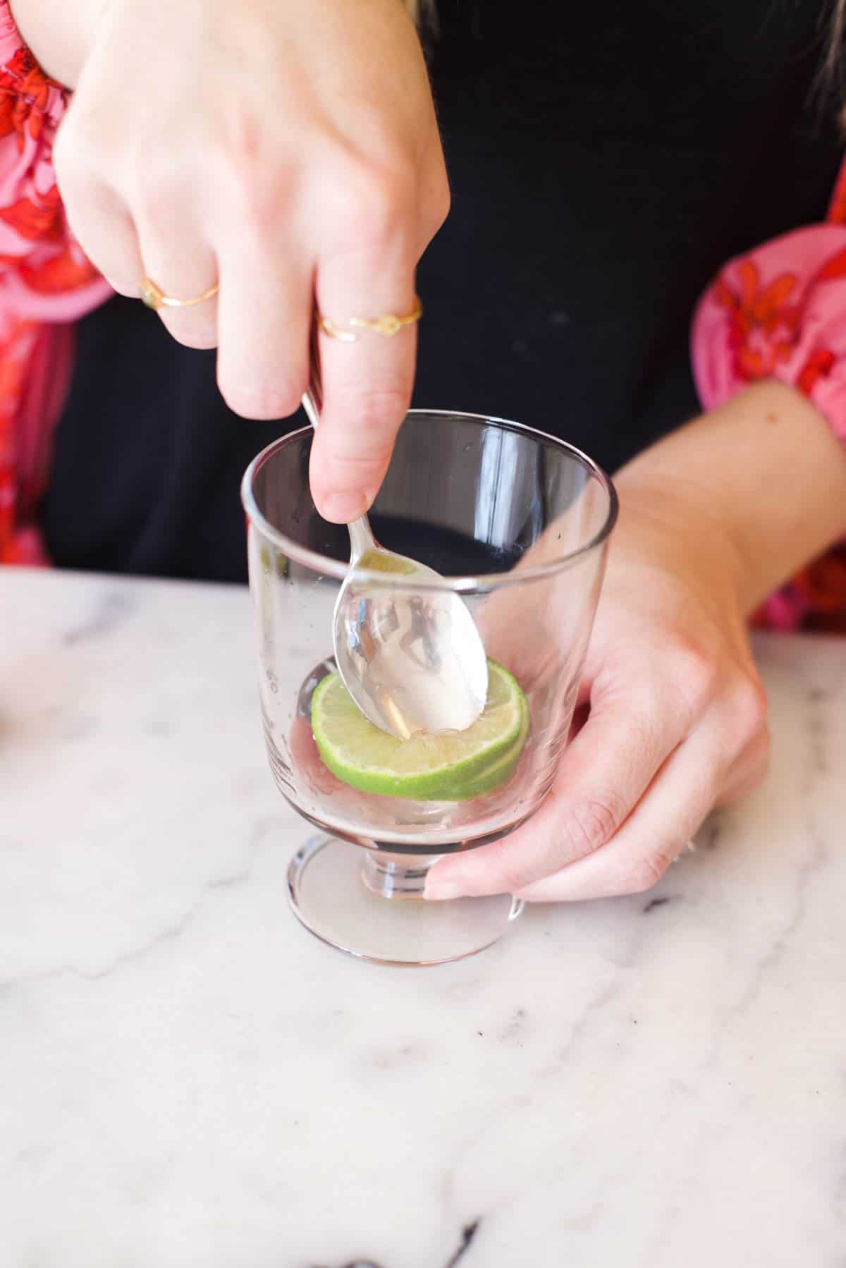 Women using a spoon to press down on a lime slice and cherries in a footed cocktail glass.