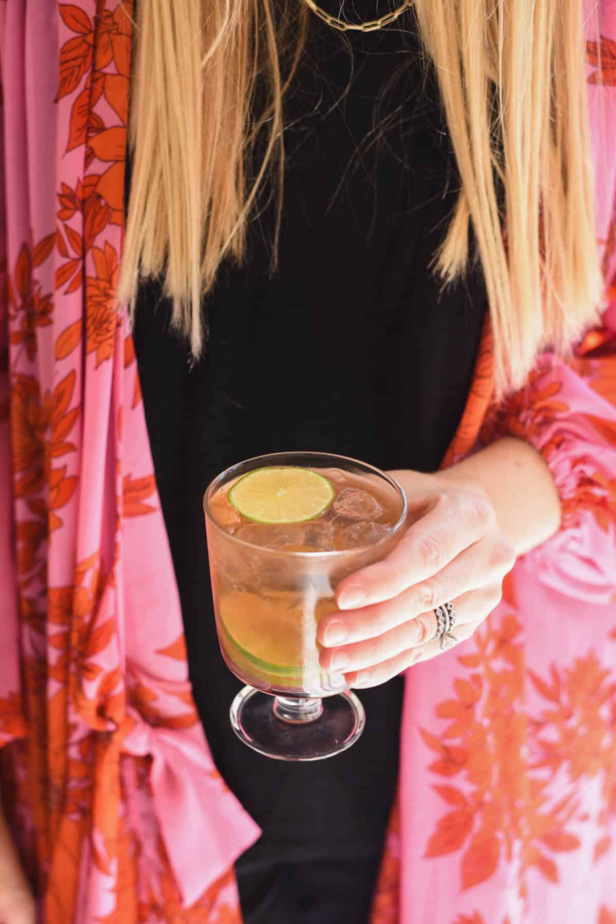 Woman holding a footed cocktail glass with a lime garnished drink.