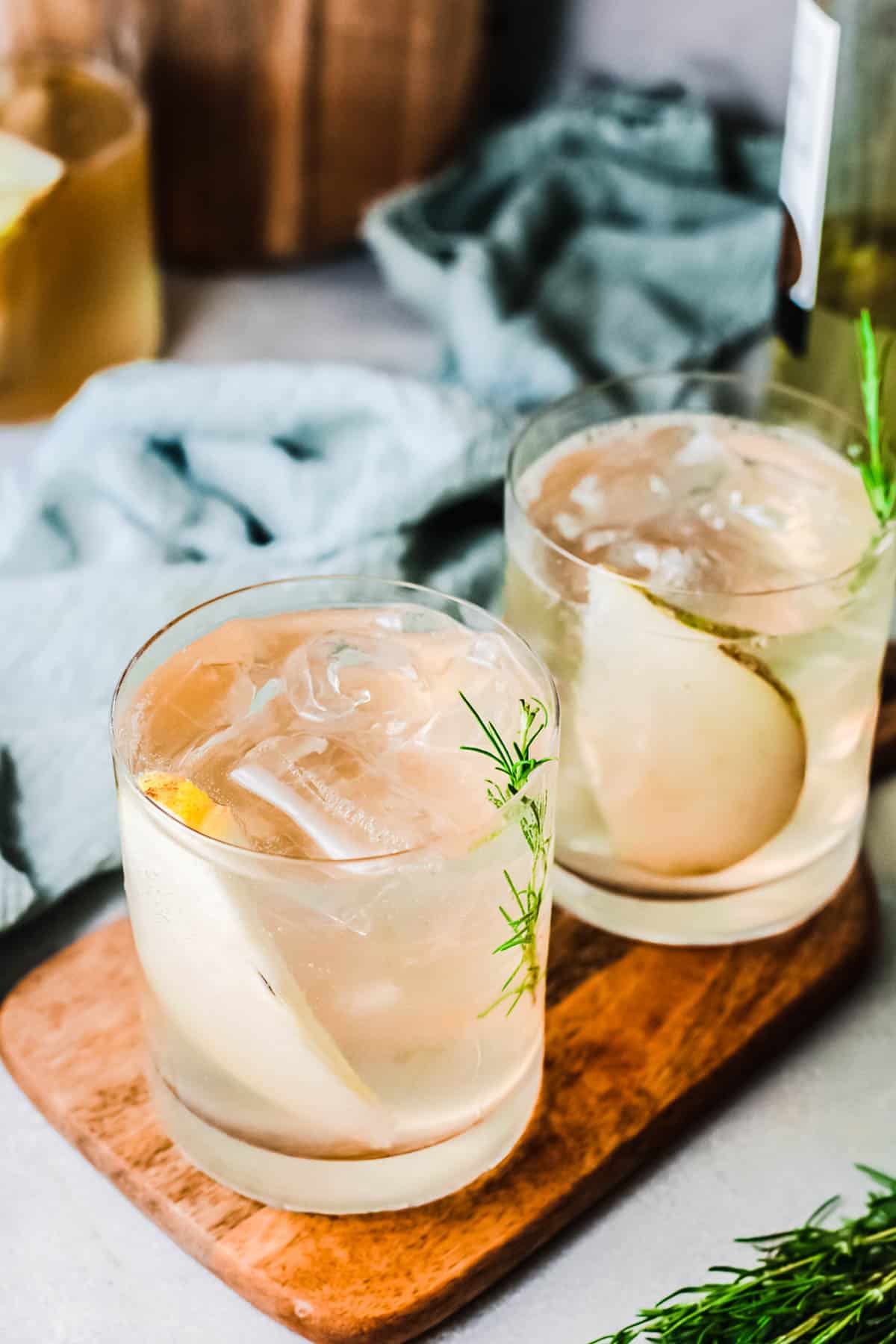 Close up of 2 cocktail glasses on a wooden tray with a sprig of rosemary and a slice of pear.