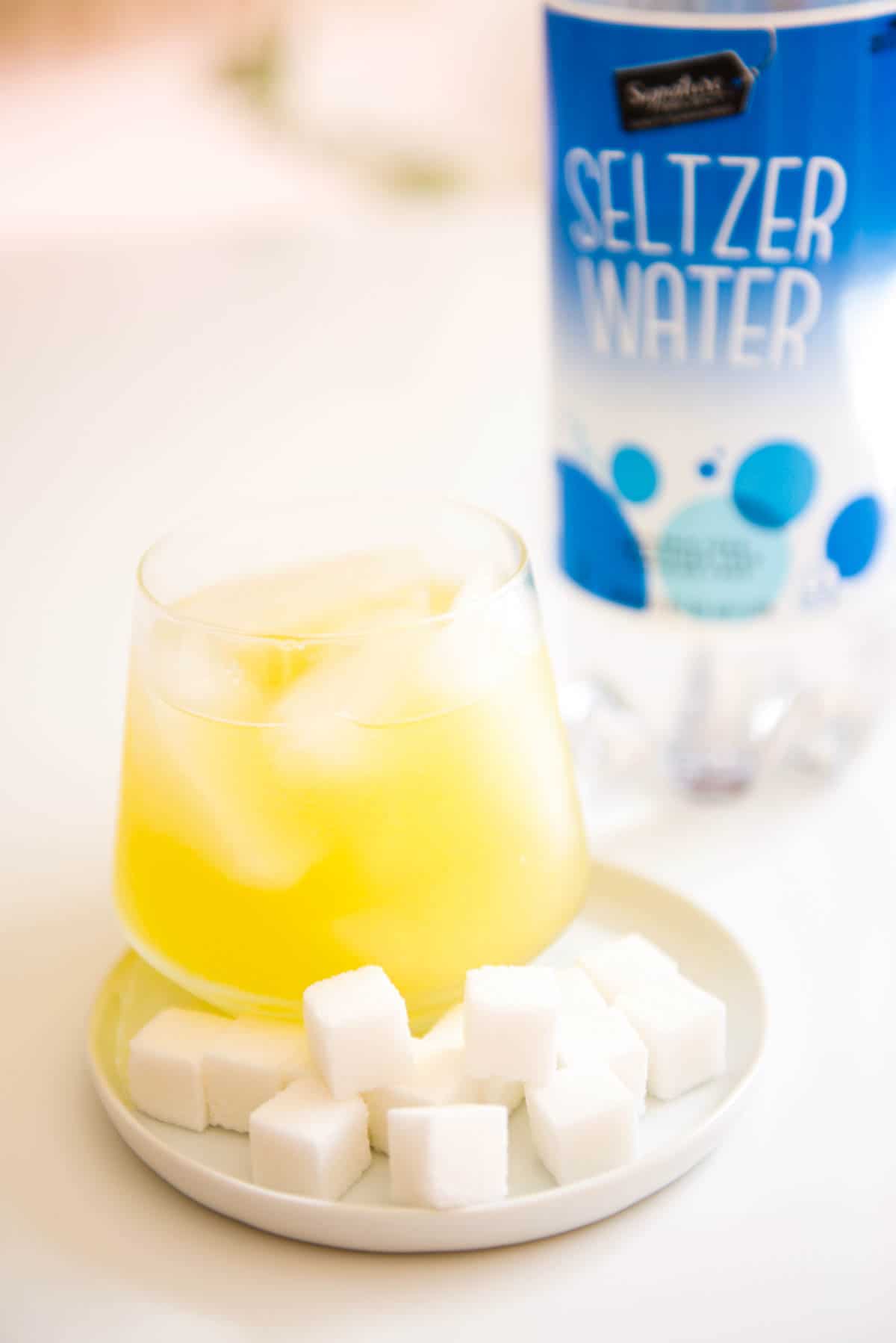 Close up of a glass holding a yellow cocktail next to sugar cubes on a plate next to a bottle of club soda.