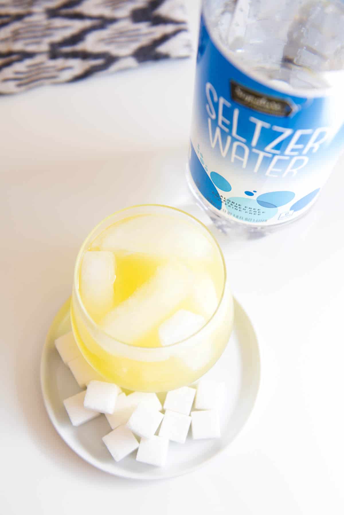 A yellow cocktail next to a bottle of club soda and sugar cubes on a counter.