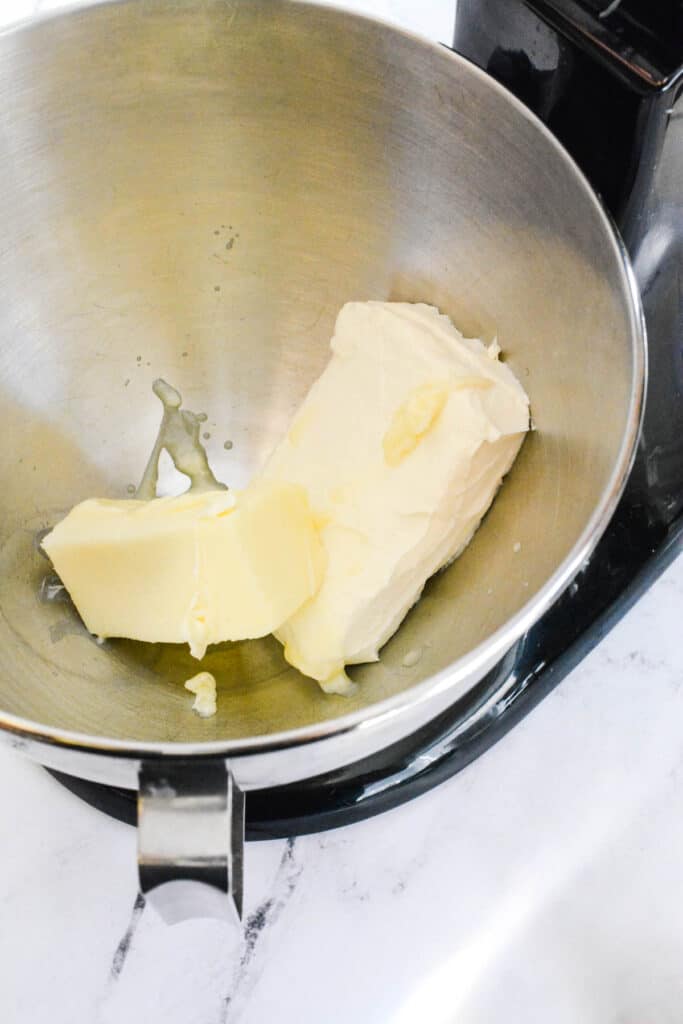 The bowl of a stand mixer with a cube of butter and brick of cream cheese.