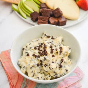 Close up of a chocolate chip cheesecake dip in a white serving bowl next to a plate of cookies and fruit.