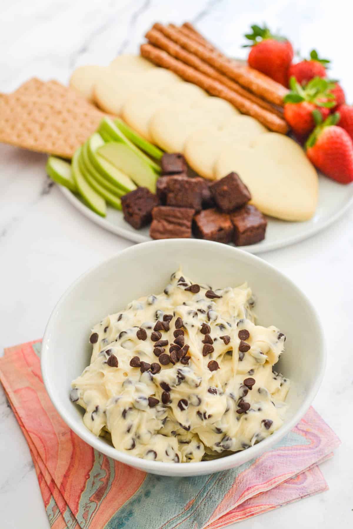 White serving bowl with cream cheese dip with chocolate chips next to a plate of dipping items like cookies.