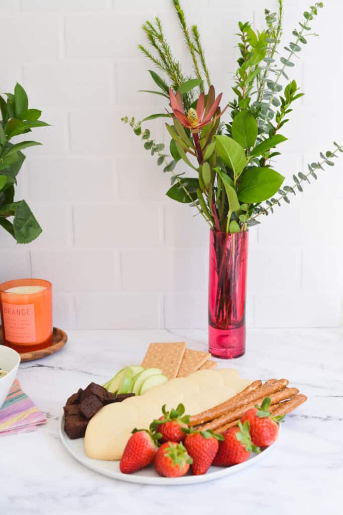 A pink vase with greenery on a table next to a plate of cookies and fruit.